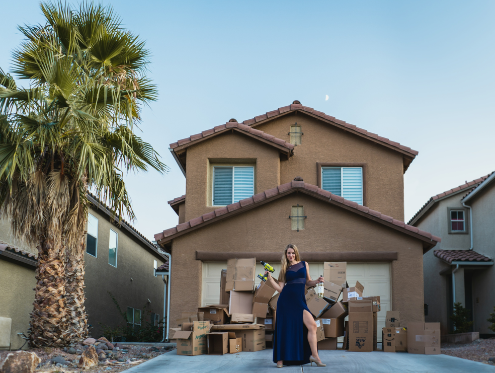 Mental Burnout After Moving House: Coping With Anxiety 2 A woman in a blue dress stands in front of a house surrounded by moving boxes, symbolizing stress and mental burnout.